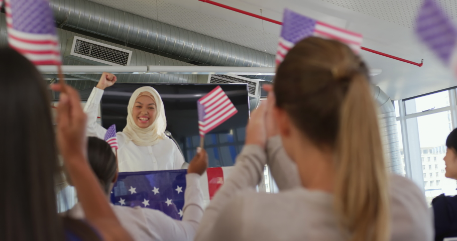 Front view of a smiling young Asian woman wearing a hijab standing at a lectern decorated with a US flag and raising her fist in triumph at a political rally, with the audience seen from the back Royalty-Free Stock Footage #1037412428