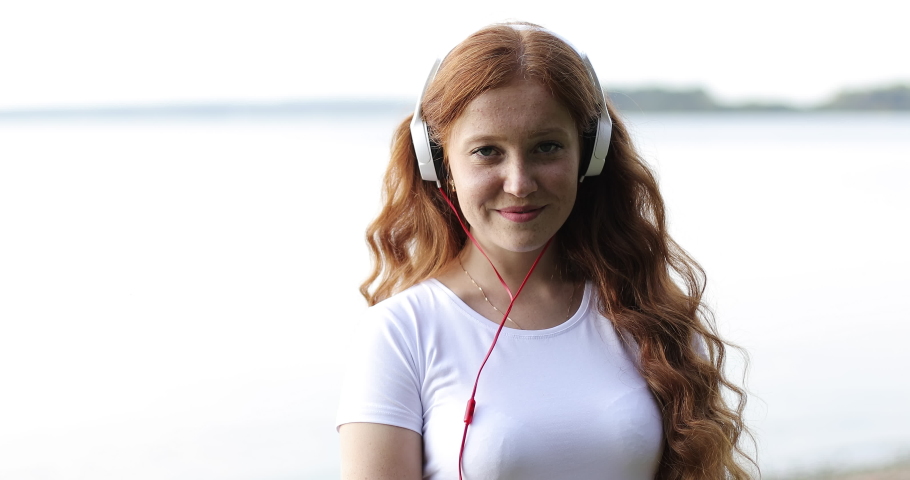 Face close up. Redhead freckled girl with white headphones is dancing on a bank of a lake listening songs. Young happy woman with red curly hair enjoying music on a shore of a lake at sunset.