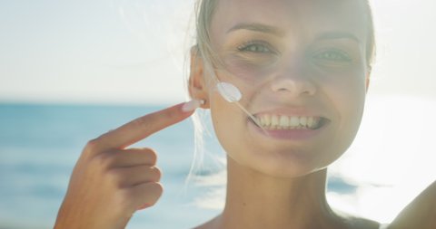 Slow motion of happy young blond hair woman is applying a sunscreen or sun tanning lotion to take care of her skin during a vacation on a beach and smiling in camera.