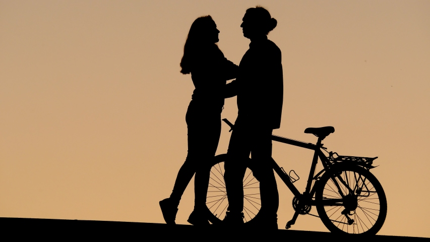 Young adult couple silhouette, man gently touch woman nose by finger. Lovely pair chatting at top against tender yellow evening sky, guy hold bicycle