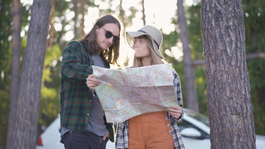 Pretty girl in hat and young man in sunglasses staying in the forest and trying to choose the way looking into the map