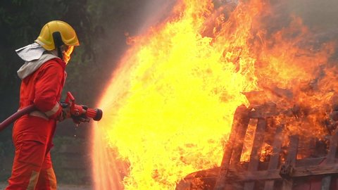 Brave firefighter in fire protection suit on safety rescue duty spray water from fire hose extinguishing crackle fire flames on explosion burning car. Fireman firefighting in danger training area