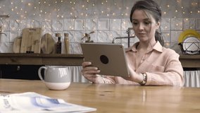 Adorable and charming dark haired woman sitting at the table against the wall with kitchen stuff and shining garland using tablet device. Stock footage. Female using modern gadget at home.