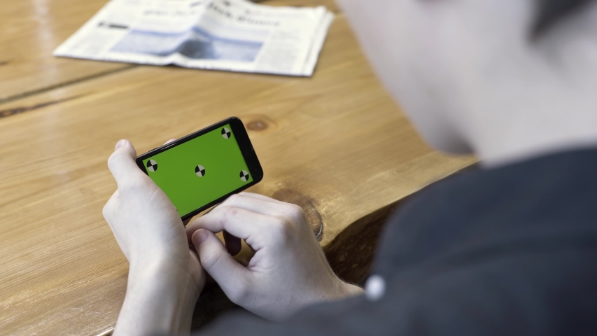 Smartphone horizontal orientation with a green screen: shot over the shoulder of a young man. Stock Footage. Man sitting at the table, tapping and scrolling on the chromakey of the phone. | Shutterstock HD Video #1037425157