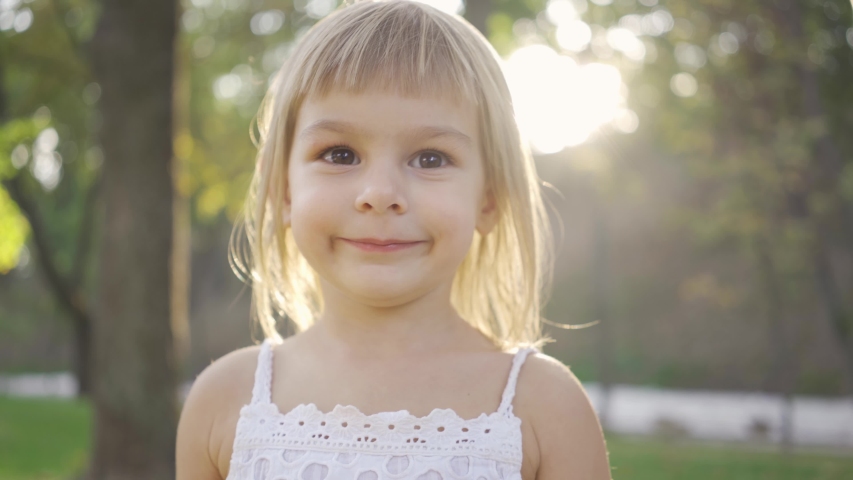 Close-up of a cute blond girl looking at the camera and laughing. Portrait of bautiful child in the park in sun rays. Leisure activity, outdoors.