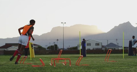 Side view of two young adult mixed race female rugby players wearing bibs jumping agility hurdles during a training session, with teammates in the background  in slow motion