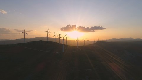 Wind turbine eco farm on beautiful golden hour evening mountain landscape. Renewable energy production for green ecological world. Aerial view of wind mills farm park, evening mountain. Lateral flight