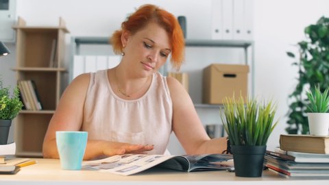 Young redhead woman in office read book with cup of tea or coffee. Relaxing on work with reading interesting books. 4k.