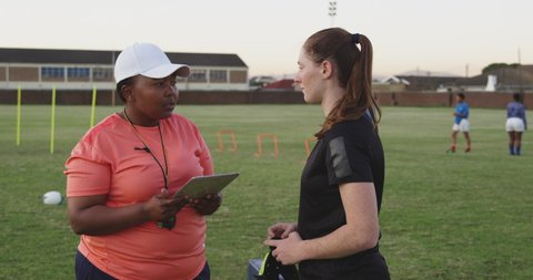 Side view of a middle aged mixed race female rugby coach holding a tablet computer talking to a young adult Caucasian female rugby player at a sports field during a training session in slow motion