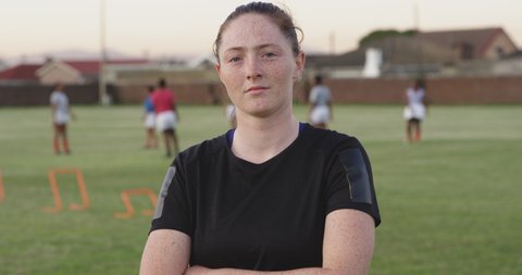 Portrait close up of a young adult Caucasian female rugby player standing on a rugby pitch looking to camera, with her teammates training in the background in slow motion