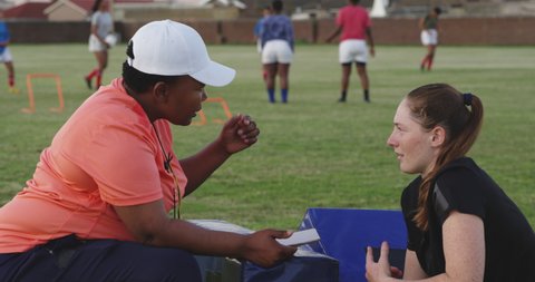 Side view of a middle aged mixed race female rugby coach talking to a young adult Caucasian female rugby player at a sports field during a training session in slow motion