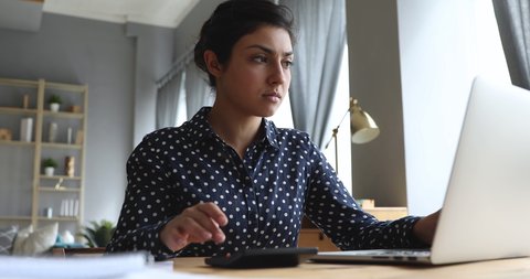 Serious young indian woman calculate domestic bills pay loan payment online on laptop sit at home office desk, focused businesswoman using computer calculator plan expenses manage finances concept