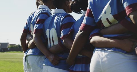 Rear view mid section of a team of young adult multi-ethnic female rugby players standing on a rugby field with arms linked, preparing for a rugby match in slow motion