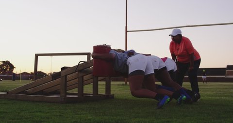 Side view of a middle aged mixed race female rugby coach instructing three young adult African American female rugby players using a scrum machine during a team training  in slow motion