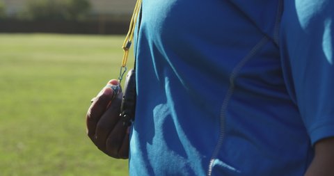 Side view close up of a middle aged mixed race female rugby coach standing on a sports field blowing a whistle in slow motion