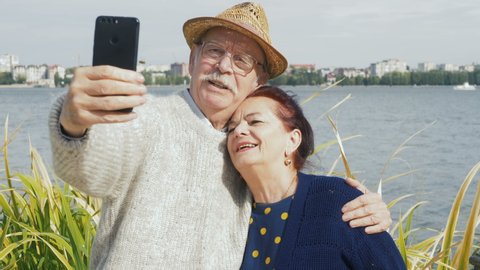 Attractive happy elderly couple in 60s take selfie with smartphone at sunset on lake smiling and laughing together. Retired baby boomers using cell phone for photos and sharing on social media apps