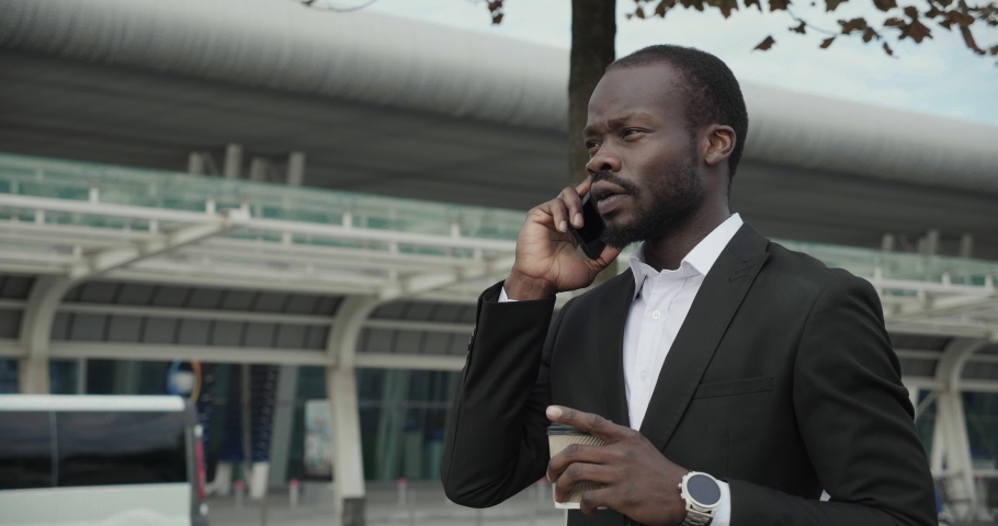Attractive Man walking in the Town while speaking on Mobile Phone. Looking Confident. Drinking Tasty Coffee from Carton cup. Many buses by the Airport waiting for tourists. Slow motion.