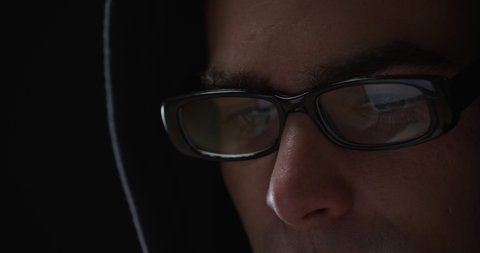 Head shot close up of a young Caucasian man wearing a black hoodie and glasses sitting, looking down and typing on a computer, the reflection of the screen visible in his glasses 