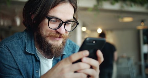 Trendy bearded student in eyewear surfing internet on smartphone and smiling, lifestyle