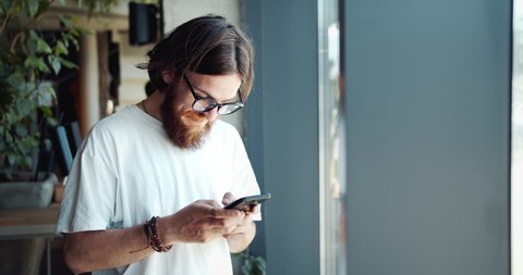 Happy smiling male hippie in eyeglasses and white t-shirt texting on smartphone in cafe, communication