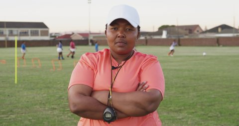 Portrait close up of a middle aged mixed race female rugby coach standing on a sports field with arms crossed looking to camera, with players training in the background in slow motion