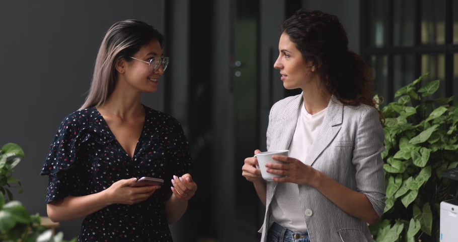 Two happy friendly young diverse business women talking during work break standing in office, caucasian and asian female employees share ideas enjoy conversation discussing team project at meeting Royalty-Free Stock Footage #1037447444