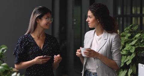 Two happy friendly young diverse business women talking during work break standing in office, caucasian and asian female employees share ideas enjoy conversation discussing team project at meeting
