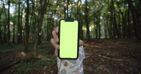 Lviv, Ukraine - 15 August, 2019: Young man showing greenscreen chrome key smartphone in hand staying in green forest on sunny day. First-person view male hand typing on mockup mobile touchscreen.