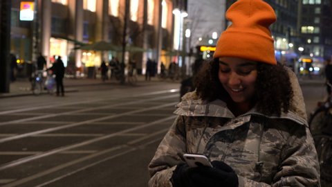 Beautiful biracial African American girl teenager young woman on urban city street at night in winter wearing orange hat and camouflage jacket using mobile cell phone smartphone for social media