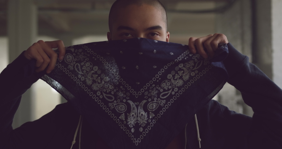 Front view of a hip young mixed race man in an empty warehouse, covering his face with bandana
