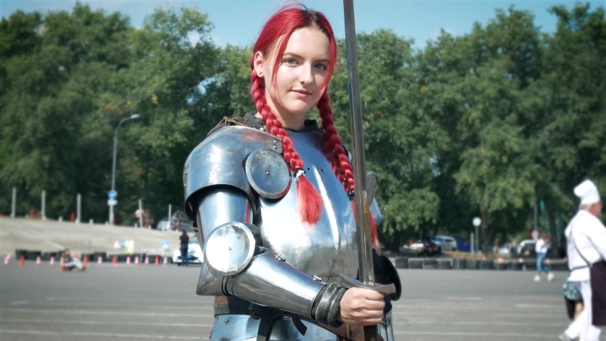 Portrait of a beautiful red haired girl in full armor set and sword. Female knight with cuirass and sword outdoors 4k