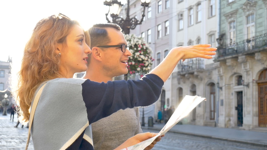 Happy young tourists couple holding a paper map of ancient European city early in the morning on empty square and pointing on some sightseeing place | Shutterstock HD Video #1037474864