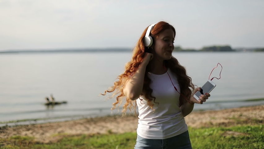 Slow motion. Young happy woman with red curly hair enjoying music and dancing in earphones on a shore of lake. Sun kissed redhead girl in white headphones is dancing near the river. Time of sunset.