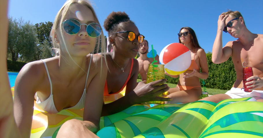Authentic shot of young multi-ethnic friends in swimwear are relaxing on colorful inflatables and making a selfie or video call with a smartphone in swimming pool in a sunny day.