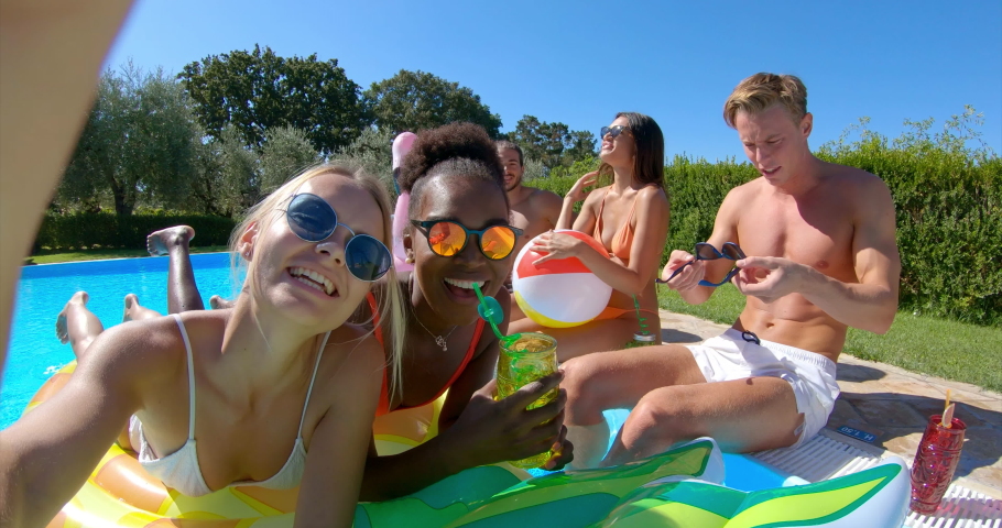 Authentic shot of young multi-ethnic friends in swimwear are relaxing on colorful inflatables and making a selfie or video call with a smartphone in swimming pool in a sunny day.