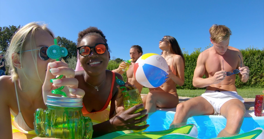 Authentic shot of young multi-ethnic friends in swimwear are relaxing on colorful inflatables and cheering with cocktails in swimming pool for celebrate a start of their vacation together.