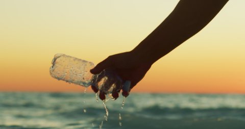 Slow motion close up of a hand of young volunteer is picking up a plastic bottle from a sea to protect an environment on a sunset.
