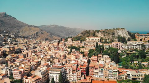 Taormina, SICILY, Italy - August 2019: Urban settlement near the rocky mountains and volcano. Red roofs of houses and people walking in the streets. Aerial drone shot.