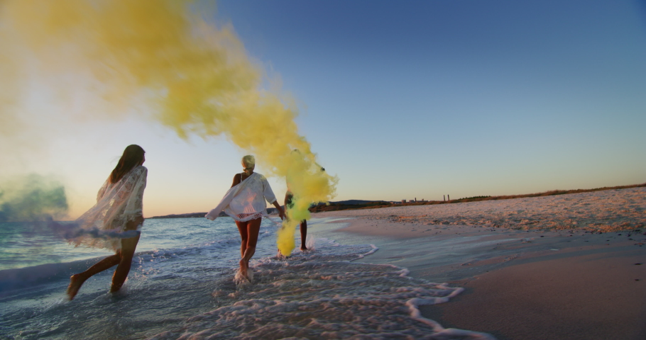 Slow motion of young multi-ethnic carefree girlfriends in swimsuits are having fun with colorful smoke flares on a beach with a sea on a sunset. Royalty-Free Stock Footage #1037482145