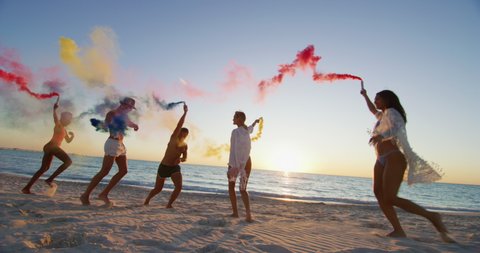 Slow motion of young multi-ethnic carefree friends in swimsuits are having fun with colorful smoke flares on a beach with a sea on a sunset.