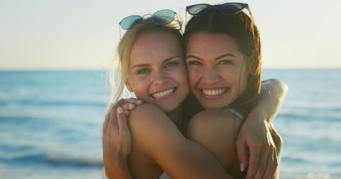 Slow motion of two young girlfriends in bikinis are having fun to enjoy their summer vacation together and embracing on a beach with a sea on a sunset.