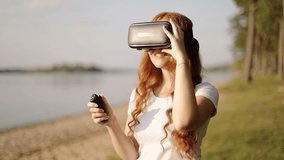 Freckled redhead girl in VR glasses with earphone is watching 360 degrees video using remote control. Virtual reality in spectacles. Slow motion, steady cam shot. 
