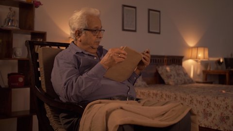 Shot of a retired Indian man reading a book sitting on a rocking chair. 4K stock video of an elderly Indian man relaxing and reading a spiritual book in his bedroom
