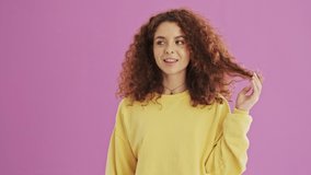 Thoughtful young redhead curly woman smiling and playing with her hair over pink background isolated