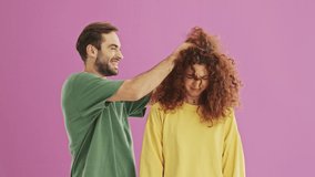 Handsome cheerful young couple smiling and playing with hair while having fun over pink background isolated