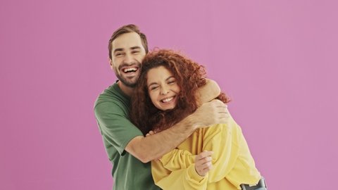 Smiling young bearded brunet man coming from the back and closing his girlfriend's eyes. Cute cheerful young lovely couple smiling and hugging over pink background isolated