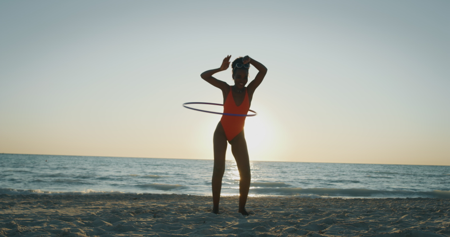 Slow motion of an young african woman in bright bikini is having fun with a hula hoop on a beach with a sea on a sunset.