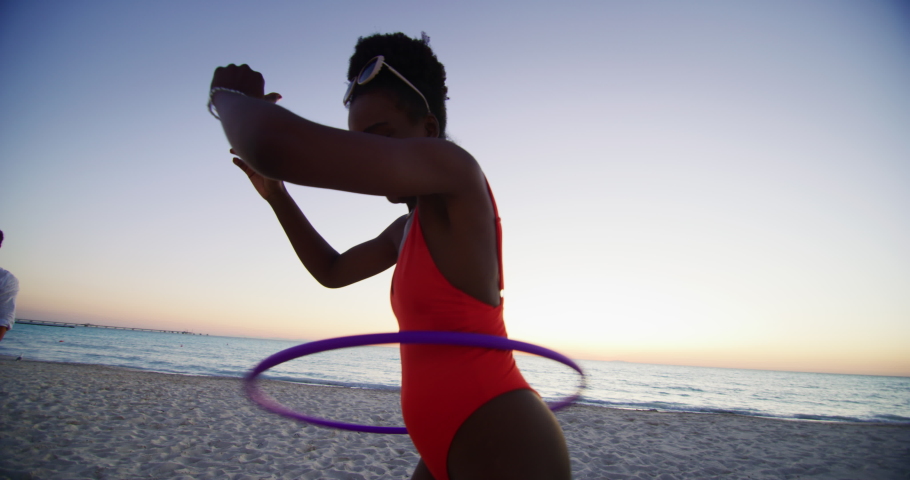 Slow motion of an young african woman in bright bikini is having fun with a hula hoop on a beach with a sea on a sunset.