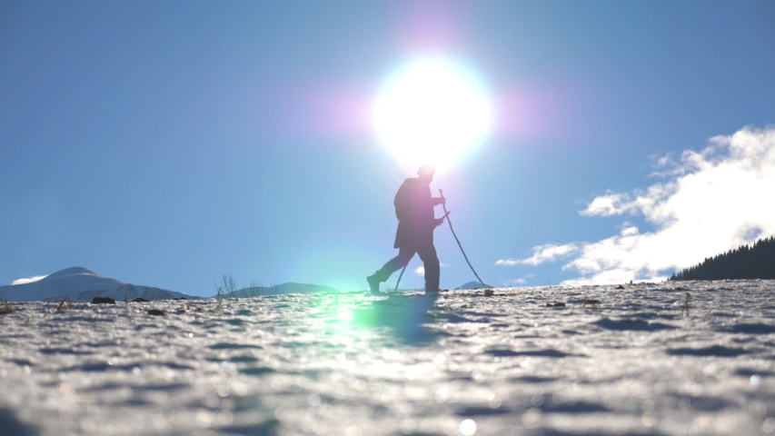 Side view of unrecognizable man going nordic walking with sticks on snowy meadow at sunny day. Young hiker trekking on snow and exercising outdoor. Concept of healthy active lifestyle. Slow motion