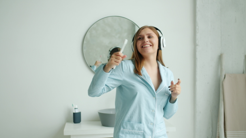 Slow motion of happy young woman in wireless headphones brushing teeth dancing enjoying music in bathroom. Modern devices, lifestyle and health concept.